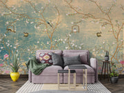 A styled living room featuring a large wall mural with a vintage-style floral branch pattern in muted teal, gold, pink, and brown tones. In front of the wall is a lavender sofa with patterned pillows and a green throw blanket, alongside two small nesting tables, potted plants, and a rug on a wood floor.