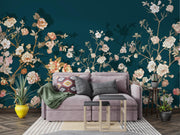 A styled living room featuring a large-scale floral wallpaper mural in deep teal with cream, pink, and peach blossoms. In front of the wall is a lavender-gray velvet sofa decorated with patterned and solid throw pillows, alongside a green throw blanket, a striped rug, and two small nesting side tables.