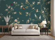 A styled living room featuring a large wall mural with a dark teal background, decorated with delicate gold branches, pink and white blossoms, green leaves, and small birds and butterflies. In front of the wall is a cream-colored sofa with metallic gold pillows, flanked by matching side tables and a lamp.