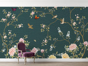 A dark teal wallpaper with a vintage Chinese floral pattern featuring pink peonies, white blossoms, gold branches, and birds, styled in a room behind a purple velvet armchair.