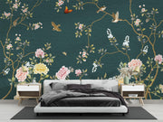A modern bedroom featuring a dark teal wallpaper with an elegant botanical pattern of pink and white flowers, green leaves, and birds in flight. A minimalist white platform bed with dark gray bedding is centered against the wall, flanked by two light wood nightstands with gold lamps.