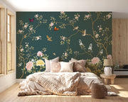 A styled bedroom featuring a large wall mural with a dark teal background, decorated with gold branches, pink and white flowers, butterflies, and birds. The room is furnished with a bed, pillows, a side table, and a lamp.