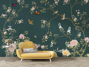 A styled interior scene showing a dark teal wallpaper with a floral and bird pattern, behind a yellow lounge chair and beside a small side table.
