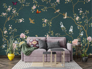 A styled living room featuring a large wall mural with a dark teal background, decorated with gold branches, pink and white flowers, birds, butterflies, and green leaves. In front of the wall is a lavender-gray sofa with patterned pillows and a green throw blanket, accompanied by two small nesting tables and potted plants.