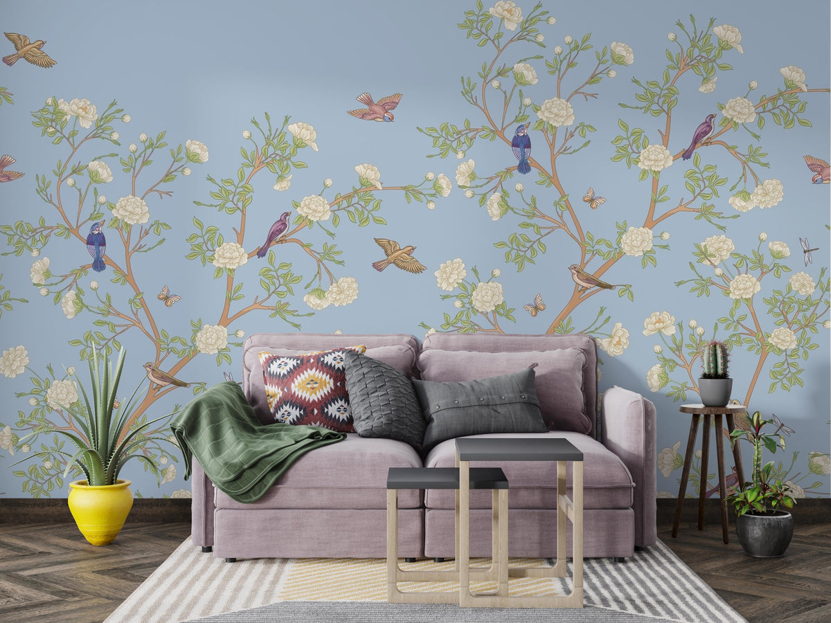 A styled living room featuring a light blue wallpaper with a detailed botanical pattern of flowering branches, birds, and butterflies. In front of the wall is a lavender sofa with patterned and solid throw pillows, a green throw blanket, and two small nesting side tables on a striped rug.