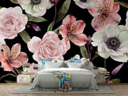 A styled bedroom featuring a large-scale floral wallpaper mural with pink roses, lilies, and white anemone flowers on a black background, behind a gray bed with colorful pillows and a patterned throw blanket.