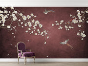 A styled interior scene showing a large wall mural with a deep burgundy background, white cherry blossom branches, and two birds, with a purple upholstered armchair placed in front of it.