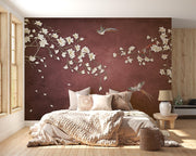 A styled bedroom featuring a large wall mural with white cherry blossom branches and birds on a deep burgundy background, behind a bed with neutral-toned bedding and pillows.