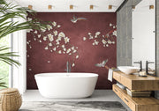 A modern bathroom interior featuring a freestanding white bathtub in front of a deep burgundy wall with white cherry blossom branches and birds. A wood vanity with a white vessel sink, gray stone wall, mirror, plant, and woven basket are also visible.