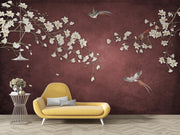 A styled interior scene with a yellow lounge chair in front of a dark burgundy wall decorated with large white blossoms and birds. A small side table with flowers sits to the left.