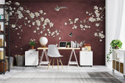 A styled home office featuring a large wall mural with white cherry blossom branches and a bird on a dark burgundy background. The room is furnished with a wooden desk, white side cabinets, a white chair, and various desk accessories.