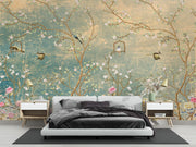 A styled bedroom featuring a large wall mural with an artistic cherry blossom tree design in soft teal, gold, pink, and white tones. The room includes a low white platform bed with dark bedding, two matching white and wood nightstands with lamps, and a dark rug on a light floor.