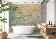A freestanding white bathtub placed in a modern bathroom with a large decorative wall mural featuring floral branches and birds in soft teal, beige, and pink tones. A wood vanity with a white vessel sink, dark faucets, gray wall panels, a mirror, and a potted plant are also visible.