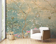 A styled interior room featuring a large wall mural with a vintage Chinese landscape design, including blossoming branches, birds, and a birdcage, in muted teal, gold, and pink tones. A white armchair and a potted plant sit in front of the wall on a light wood floor.