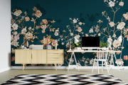 A styled home office interior featuring a dark teal floral wallpaper mural as the main product. The room includes a light wood sideboard, a white desk with a laptop, a white chair, a black and white chevron rug, and decorative plants.