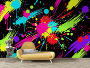 A vibrant multicolor neon paint splatter and drip pattern on a black background, styled as a wall mural behind a yellow modern lounge chair and a small side table.
