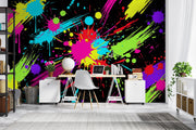 A vibrant multicolor paint splatter wall mural displayed in a modern home office setting, featuring a desk, chair, and shelving against a black background with bright neon splashes of pink, blue, green, yellow, and purple.