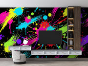 A modern living room setup featuring a bold, vibrant multicolor paint splatter wallpaper on the wall behind a floating white TV console and a black television. The wallpaper has a black base with energetic splashes of neon pink, yellow, blue, green, and purple.