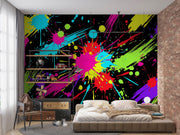 A colorful abstract wall mural with neon paint splatters and brushstrokes in black, pink, blue, yellow, green, and purple, shown behind a bed in a modern bedroom with a brick wall and shelving.