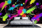 A modern bedroom setup featuring a bold, vibrant multicolor paint splatter wall mural on a black background, with a gray bed, white pillows, and matching side tables.