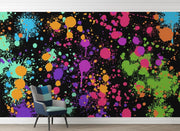 A colorful abstract paint splatter wall mural with vibrant splashes of pink, purple, green, orange, and blue on a black background, shown in a room setting with a modern armchair and light wood floor.
