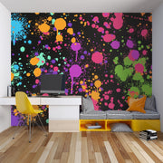 A modern home office or playroom setup featuring a bold, colorful abstract paint splatter wallpaper on the back wall. The design has a black base with vibrant splashes of pink, purple, orange, green, and blue.