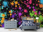 A colorful bedroom scene featuring a bold black wall mural covered in bright paint splatters and drips in pink, purple, blue, green, orange, and yellow. In front of the wall is a gray bed with plaid bedding, a matching gray dresser, a small side table with a lamp, and a skateboard on the floor.