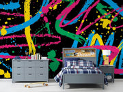 A styled bedroom featuring a bold, vibrant multicolor paint splatter and brushstroke wallpaper on a black background. The room is furnished with a gray bed frame, matching gray dresser, a gray nightstand, and a plaid blue and white comforter.