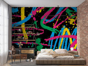 A colorful abstract wall mural with bold paint strokes in blue, pink, green, yellow, and orange on a black background, shown in a modern bedroom setting behind a bed and next to a brick wall.