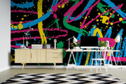 A vibrant abstract wall mural with bold neon paint splatters and brushstrokes in pink, blue, yellow, green, and orange on a black background, shown in a modern home office setting behind a light wood sideboard, white desk, and black-and-white chevron rug.