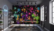 A modern office or study room with a large wall mural featuring a vibrant multicolor paint splatter pattern on a black background. The room includes a wooden desk, a green chair, shelving, and windows.