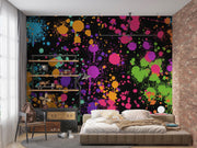 A colorful spray paint splatter wall mural on a bedroom wall, featuring vibrant pink, purple, green, orange, and blue paint splashes on a black background, styled with a bed, shelving, and brick wall.