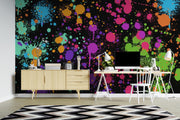 A vibrant multicolor paint splatter wallpaper mural on a wall in a modern home office setting, featuring a light wood credenza, a white desk with a computer, and a black and white chevron rug.