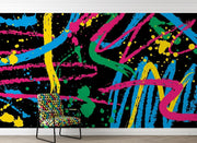 A colorful abstract wall mural with neon paint splatters and brushstrokes in blue, pink, yellow, green, and orange on a black background, shown in a room setting behind a patterned armchair.