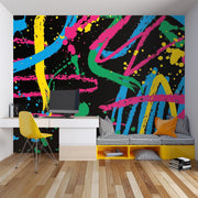 A colorful abstract wall mural with neon paint splatters and brushstrokes in pink, blue, yellow, green, and orange on a black background, shown in a modern room setting with a desk, chair, and bench.
