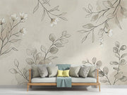 A styled interior scene showing a light wood sofa with gray and yellow pillows and a teal throw, set against a large wall mural with delicate botanical leaf and blossom illustrations on a warm beige background.