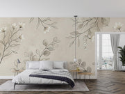 A styled bedroom interior featuring a large wall mural with delicate beige and gray botanical leaf patterns on a warm cream background. The room includes a bed with white bedding, two small wooden side tables, a hanging pendant light, and a window looking out onto a city view.