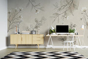 A styled home office interior featuring a large-scale floral wallpaper with delicate gray and white botanical illustrations on a warm beige background. The room is furnished with a light wood sideboard, a white desk with a computer setup, a white chair, and a black, white, and beige chevron-patterned rug.