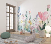 A children's playroom corner featuring a large watercolor-style floral wall mural with pink tulips, blue flowers, and green stems. The room includes a light wood floor, a gray patterned rug, a green pouf, a gray bunny-shaped bean bag, a white floor lamp, and a window with wooden frames.