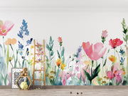 A colorful watercolor floral wall mural featuring pink, blue, yellow, and orange flowers with green stems, displayed on a white wall in a styled nursery room with a wooden floor, a wooden ladder, and a small framed sign.