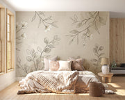 A styled bedroom interior featuring a large floral and botanical wall mural with beige and gray leaves and white blossoms behind a bed. The room has light wood flooring, a window on the left, a bedside table with a lamp, and a woven basket on the floor.
