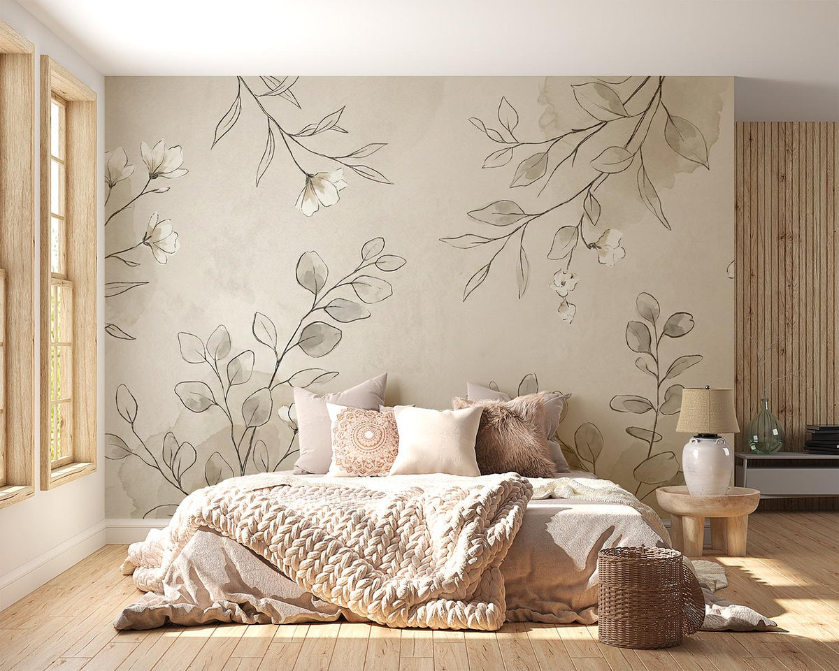 A styled bedroom interior featuring a large floral and botanical wall mural with beige and gray leaves and white blossoms behind a bed. The room has light wood flooring, a window on the left, a bedside table with a lamp, and a woven basket on the floor.