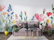 A styled living room featuring a large watercolor floral wall mural with pink, blue, yellow, and orange flowers on a white background. A lavender-gray fabric sofa sits in front of the wall, decorated with patterned and solid throw pillows, a green blanket, and a striped rug.