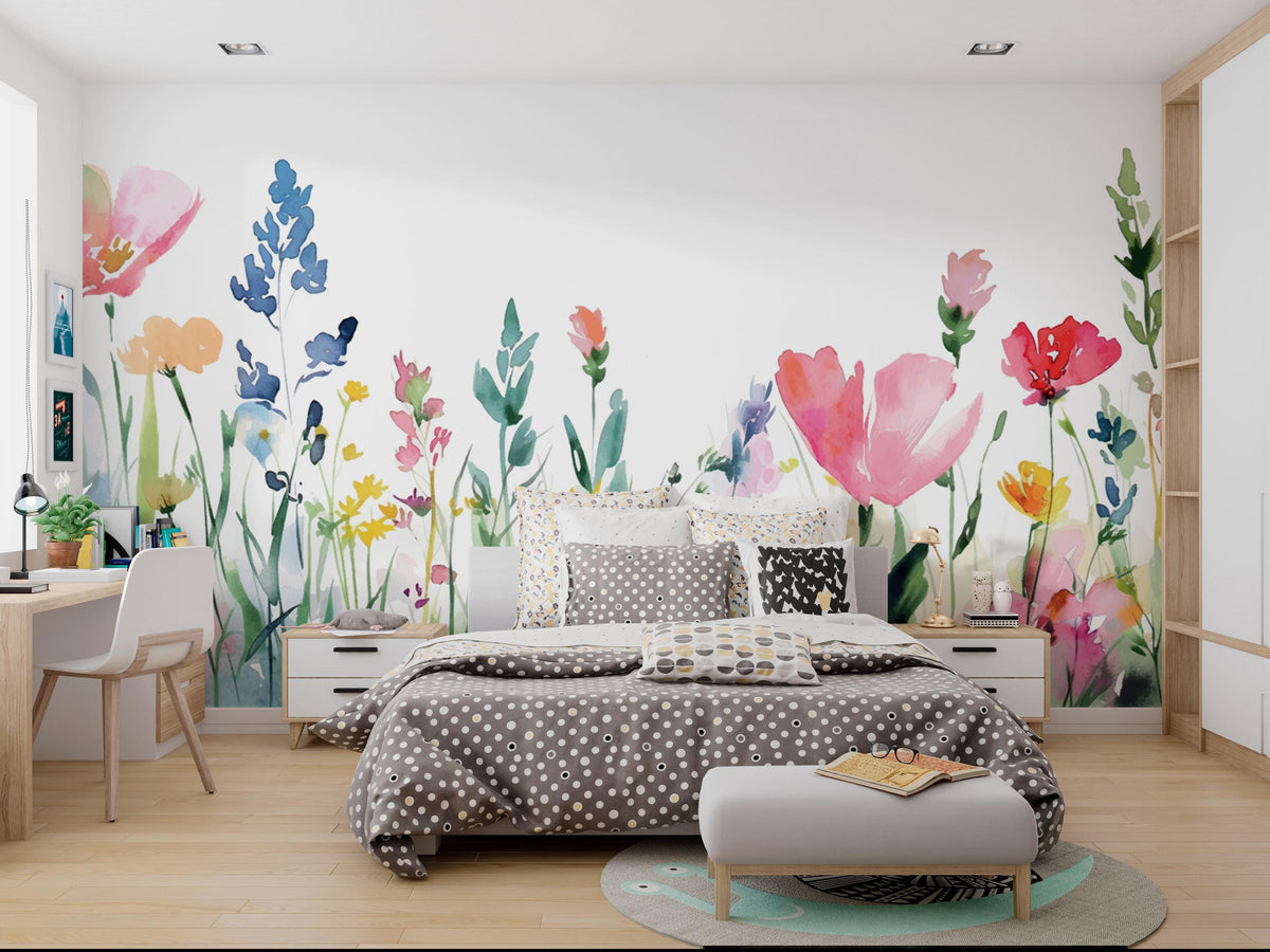 A modern bedroom featuring a large watercolor floral wall mural with pink tulips, blue flowers, and green leaves on a white wall. The room is styled with a gray and white patterned bedspread, matching pillows, a white bench, a wooden desk, and light wood flooring.