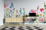 A styled home office interior featuring a large floral watercolor wall mural with pink, blue, and yellow flowers. In front of the wall is a light wood sideboard, a white desk with a computer monitor, chair, and small plants, and a black, white, and gray chevron rug on a light wood floor.