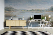 A large wall mural depicting a serene lake landscape with reeds, trees, and birds, shown in a styled home office setting with a light wood sideboard, desk, chair, and a black-and-white chevron rug.