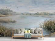 A large wall mural depicting a serene lake landscape with reeds, distant trees, flying birds, and a cloudy sky, shown behind a light wood sofa with neutral and yellow pillows in a staged interior setting.