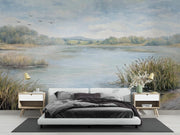 A large-scale wall mural depicting a serene lake landscape with grassy reeds, distant trees, and a cloudy sky, shown installed behind a modern white bed in a styled bedroom setting.