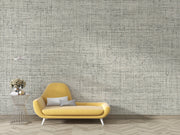 A modern yellow chaise lounge chair with two gray pillows, set against a wall covered in textured gray and white script-patterned wallpaper. A small wire side table with a vase of flowers sits to the left, and light wood herringbone flooring is visible below.