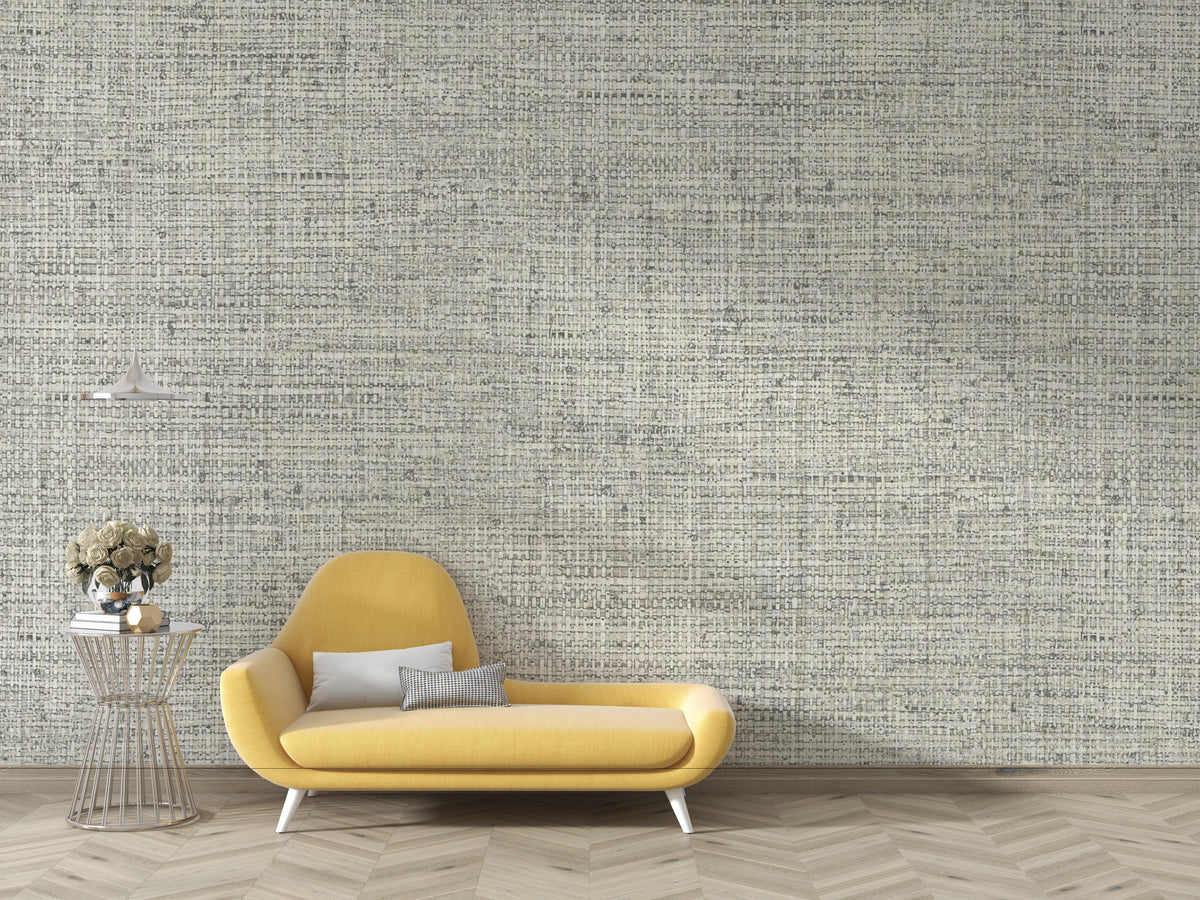 A modern yellow chaise lounge chair with two gray pillows, set against a wall covered in textured gray and white script-patterned wallpaper. A small wire side table with a vase of flowers sits to the left, and light wood herringbone flooring is visible below.